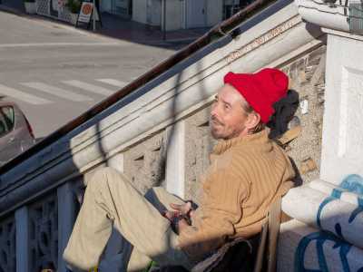 Quando lo incontriamo � seduto sui gradini, indossa un cappello rosso sui capelli biondi ed � intento a parlare con un passante